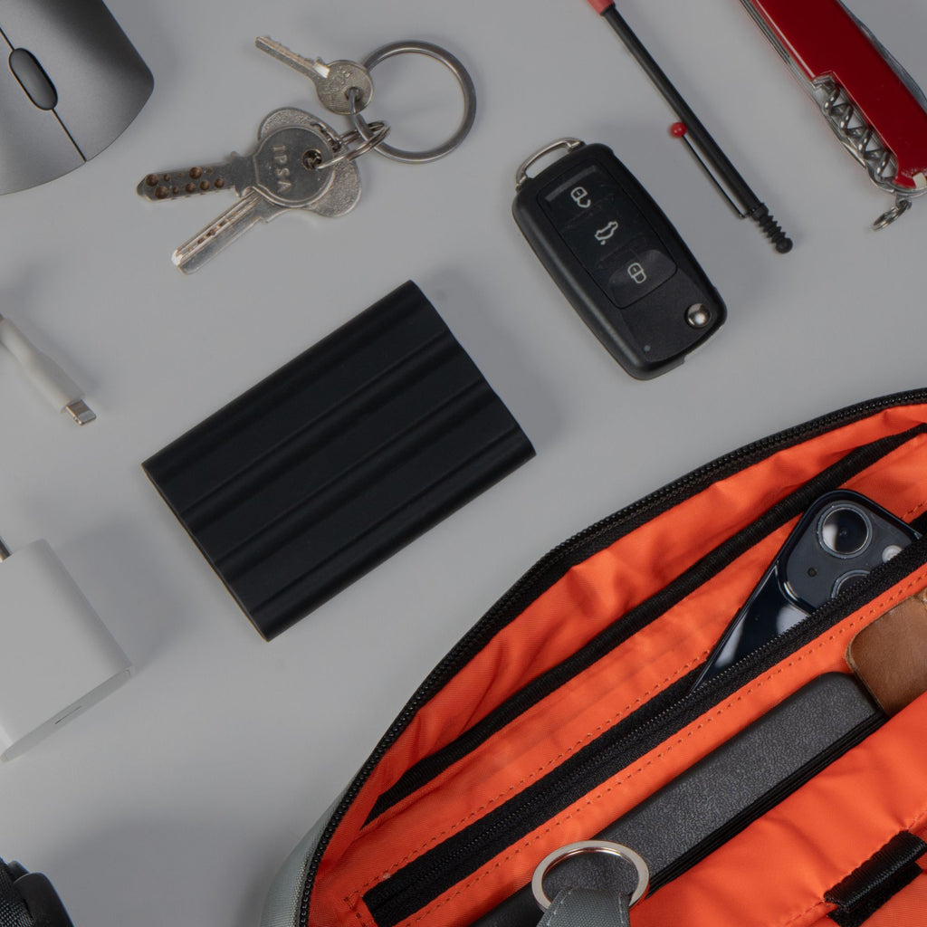 Everyday carry gear arranged next to the open orange interior pocket of the black sling bag.